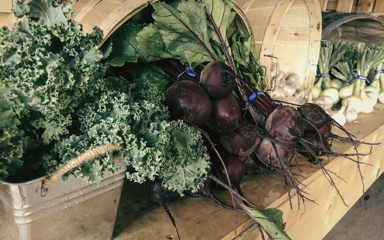 Bunches of beets and curly kale displayed in baskets on a wooden farm stand table